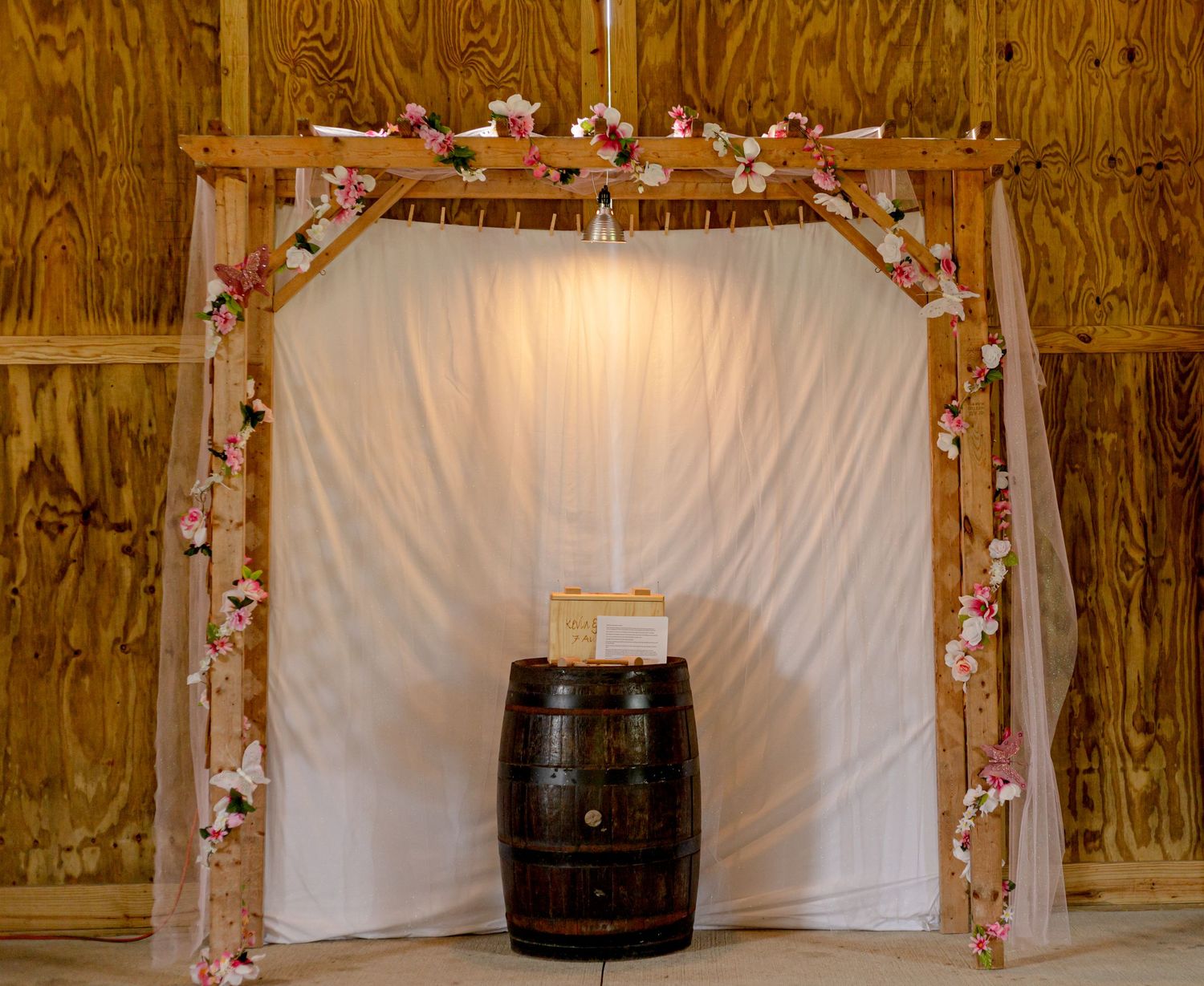 Rustic barn wedding arch decorated with pink flowers and white draping over wine barrel.