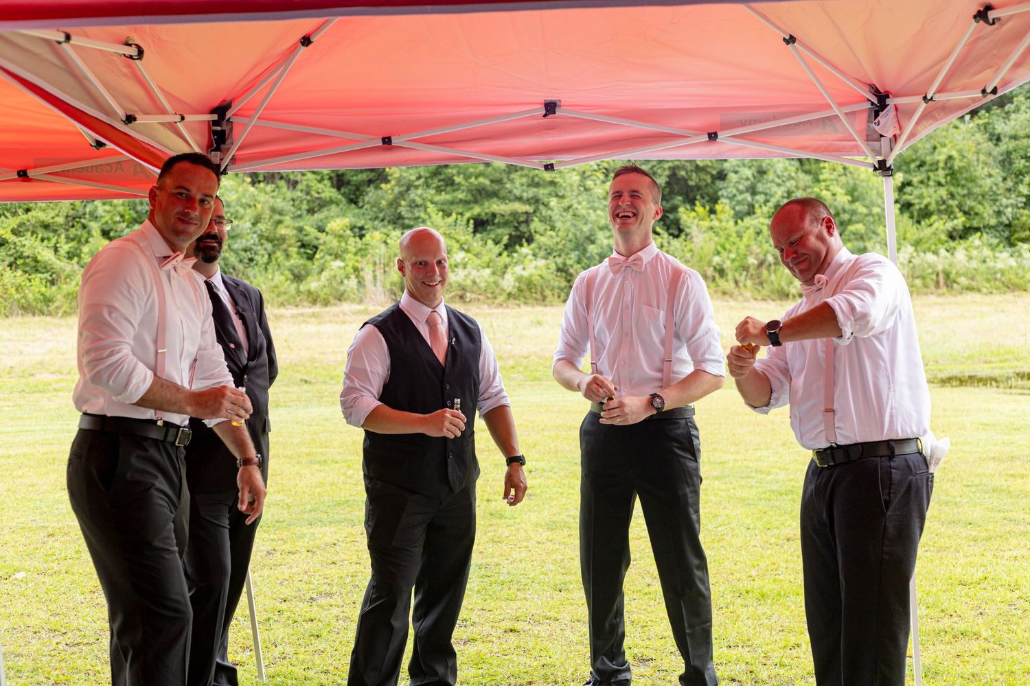 Four groomsmen in white shirts and black pants laughing under a red outdoor canopy.