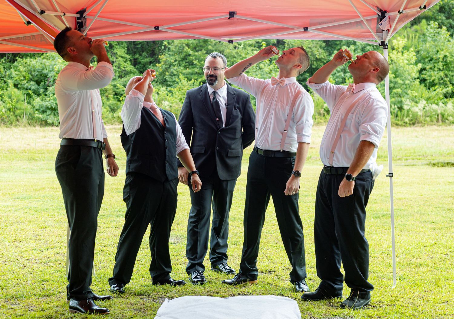 Five groomsmen in white shirts toasting under a red canopy at an outdoor wedding.