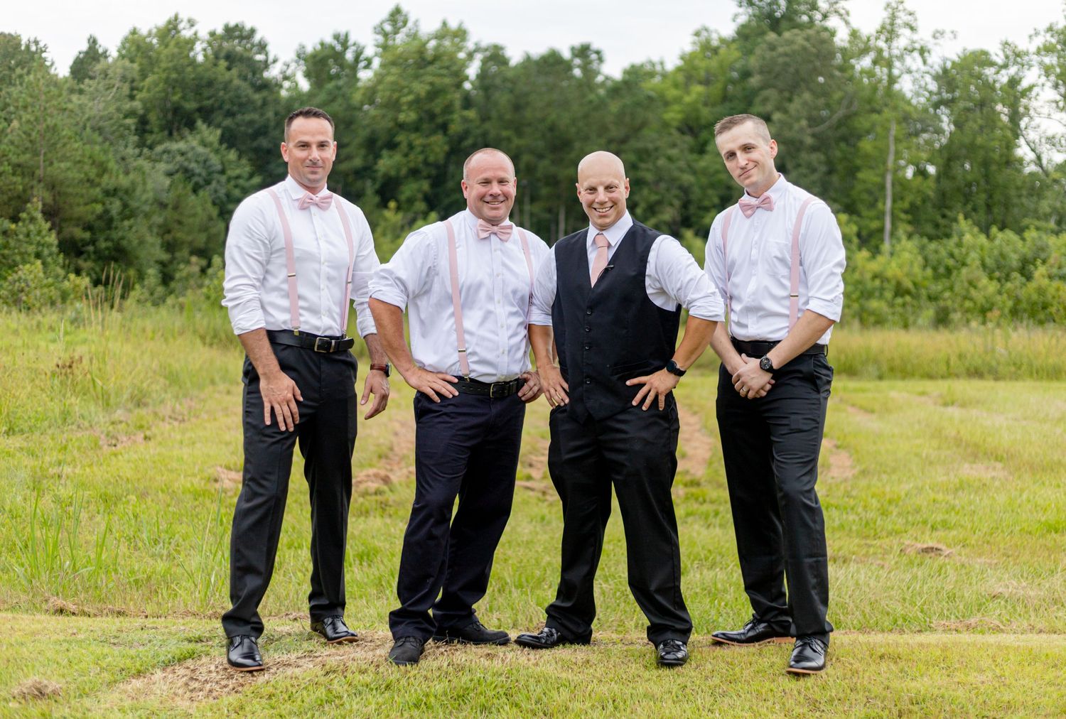 Four groomsmen in white shirts and black pants posing in a green outdoor field.