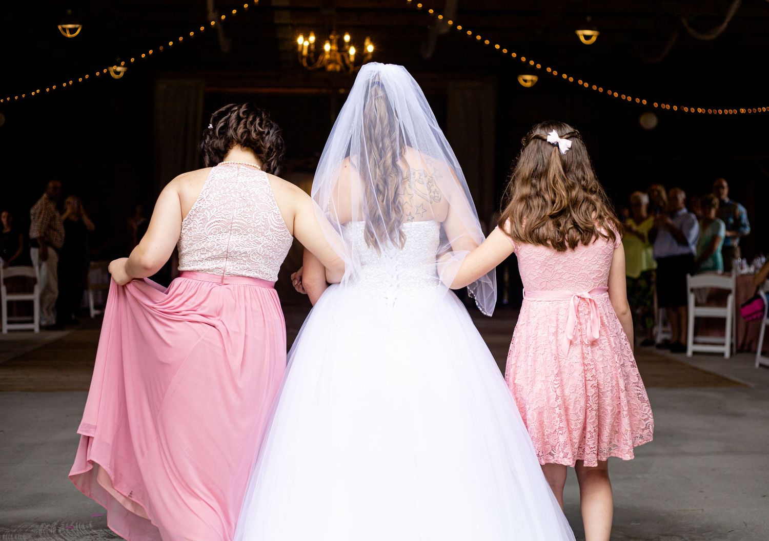 Bride in white gown and veil walking with bridesmaids in pink dresses from behind.