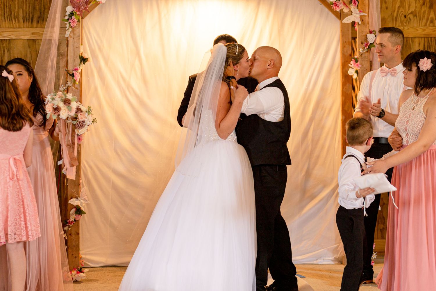 Bride and groom sharing first kiss under floral arch at rustic barn wedding ceremony.