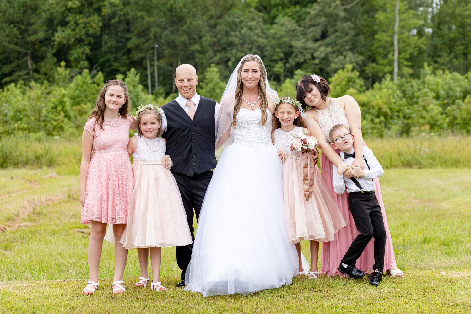 Bride and groom posing outdoors with flower girls and ring bearer in pink attire.