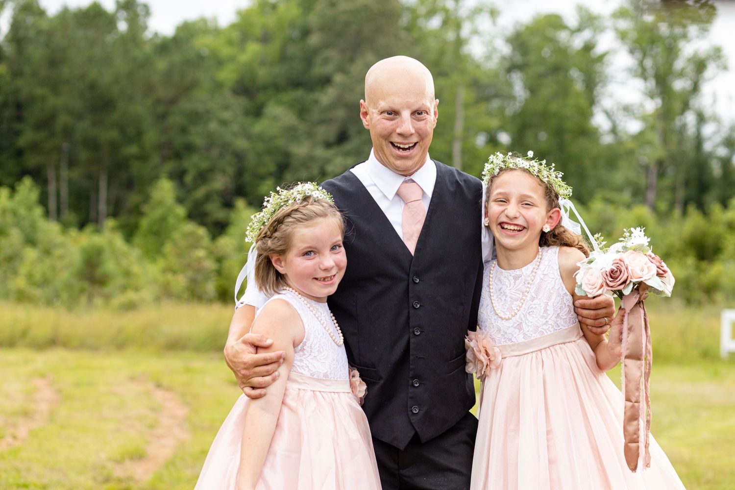 Bald groom smiling with two flower girls in blush dresses and floral crowns at outdoor wedding.