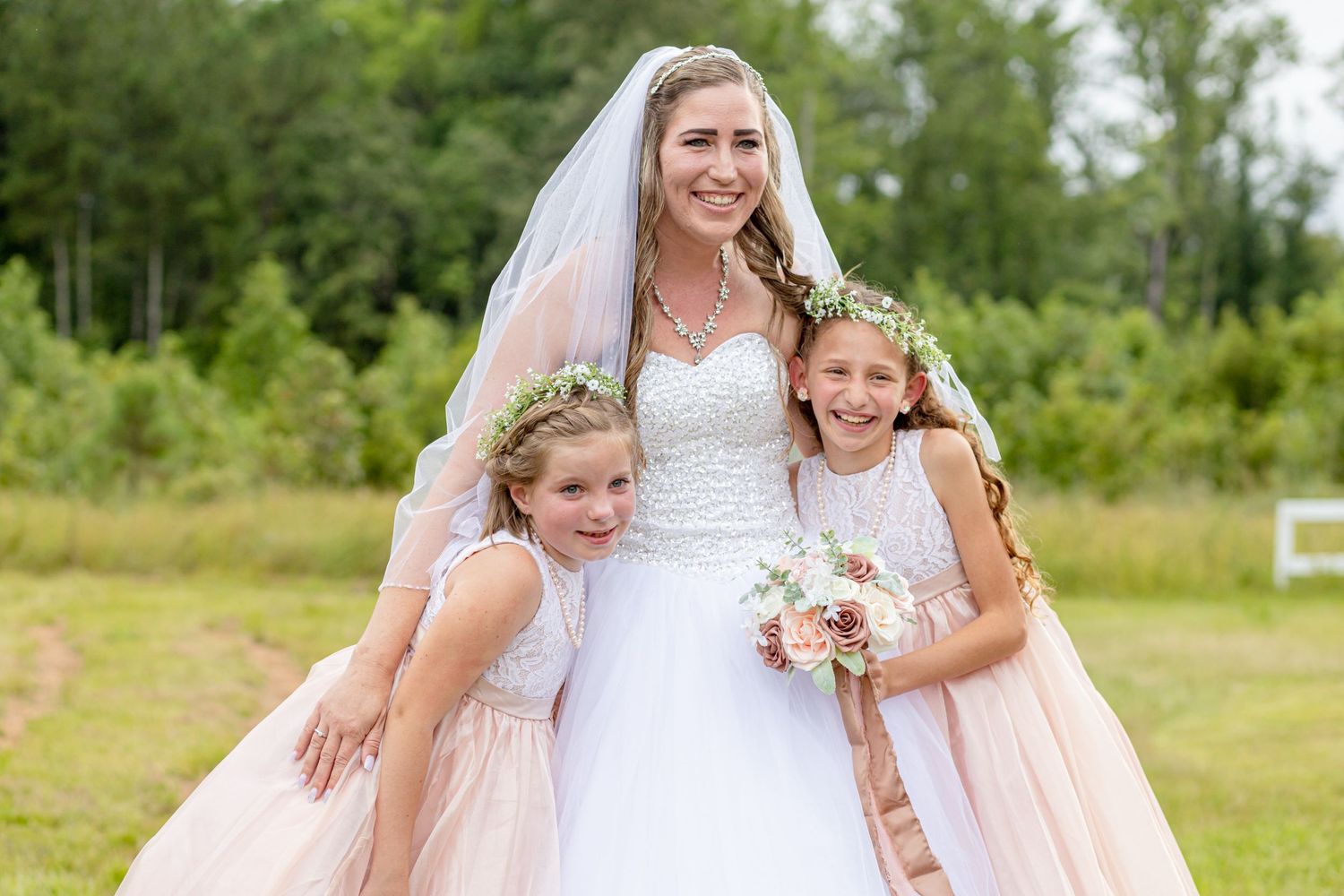 Smiling bride in white gown with veil posing with two flower girls in blush dresses outdoors.