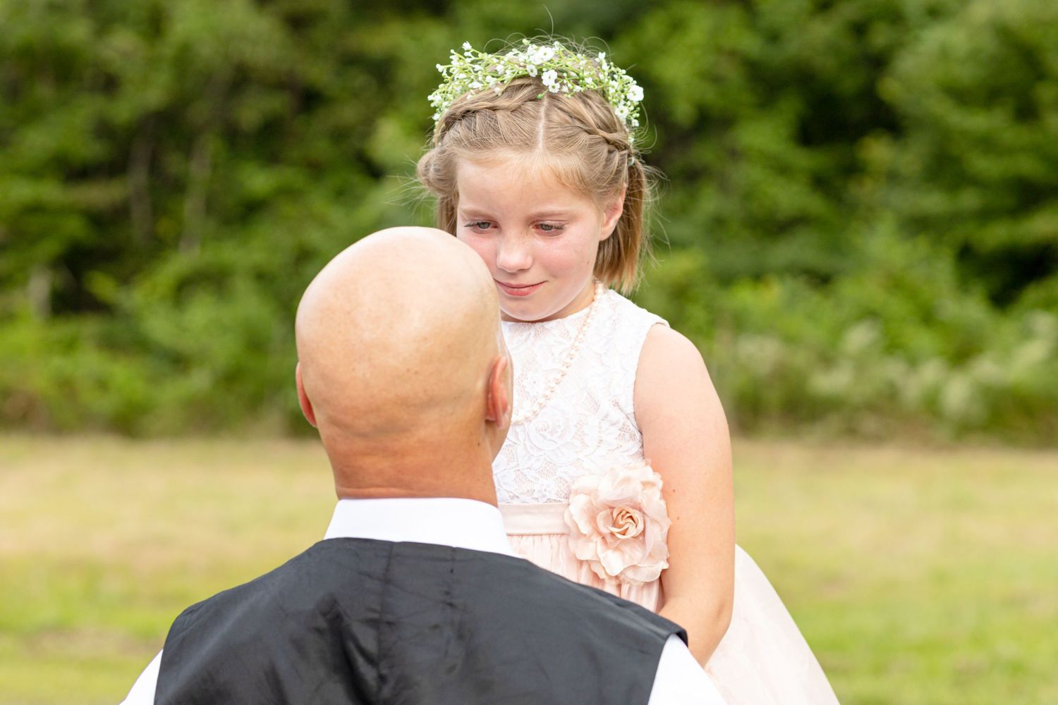 Flower girl in white lace dress and floral crown gazing at bald groom during outdoor wedding.