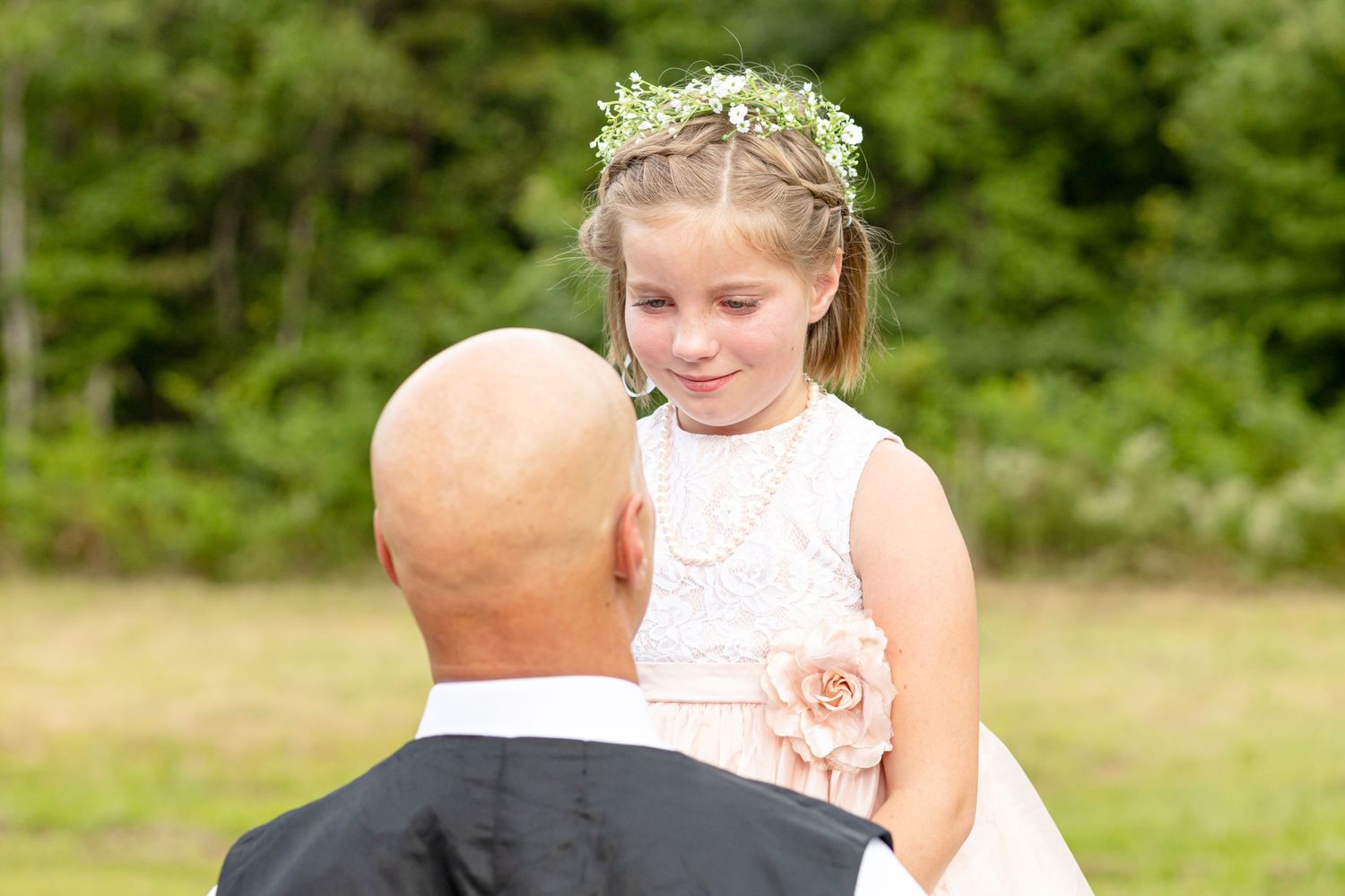 Young flower girl in white lace dress with floral crown looking at bald groom outdoors.