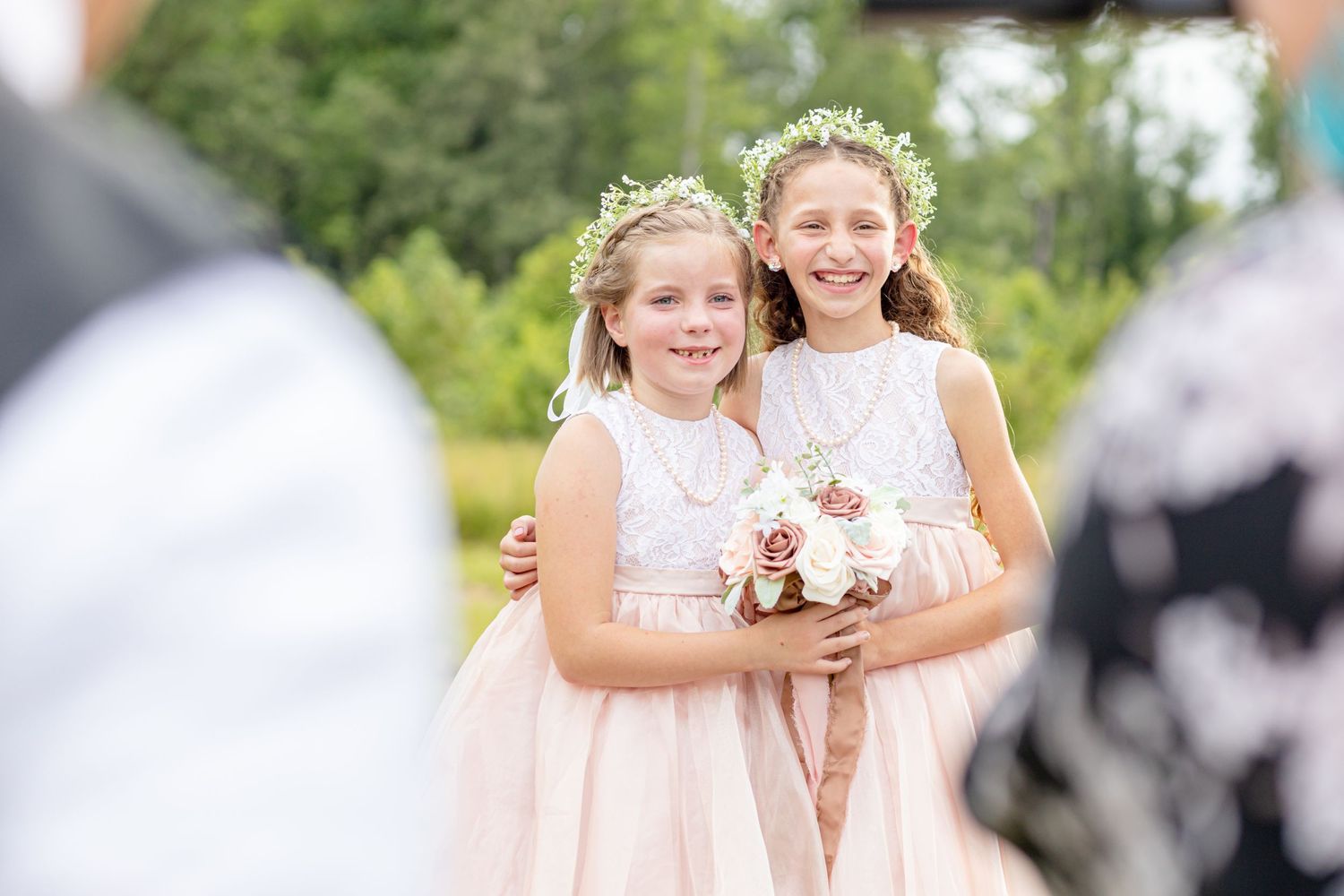 Two flower girls in matching blush dresses and floral crowns holding bouquet at outdoor wedding.