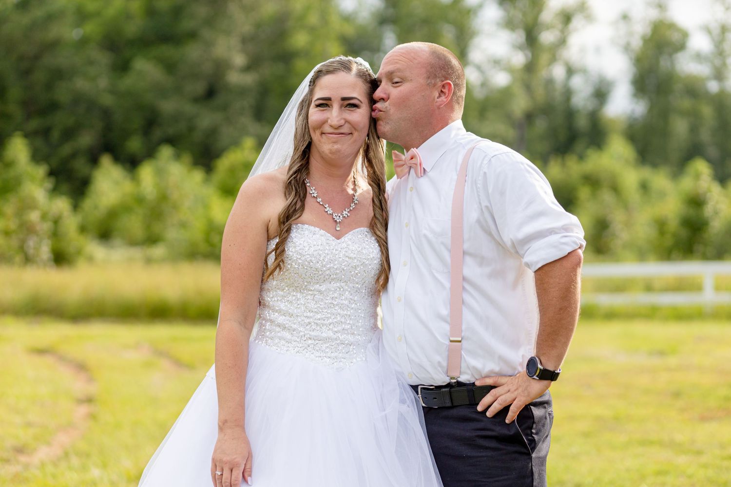 Groom in white shirt with pink suspenders kissing bride in white wedding gown outdoors.