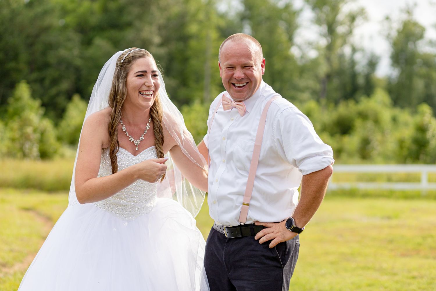 Laughing bride and groom in white shirt with pink suspenders sharing a joyful moment outdoors.