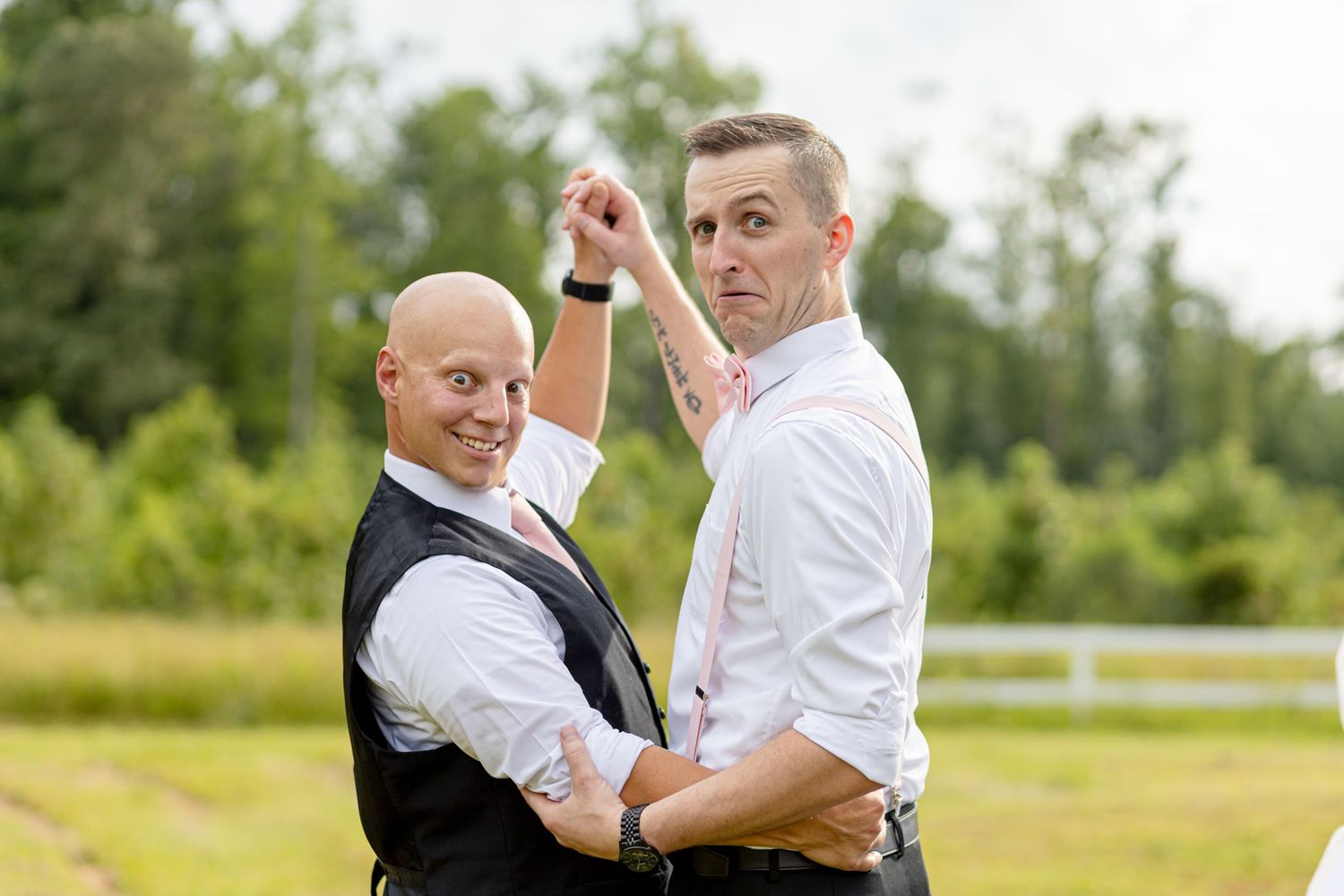Two men in wedding attire playfully posing together in a field at an outdoor wedding.