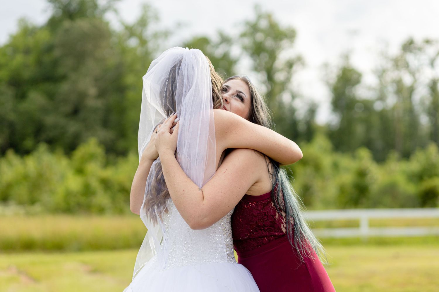 Bride in white gown and veil sharing a warm embrace with woman in burgundy dress outdoors.