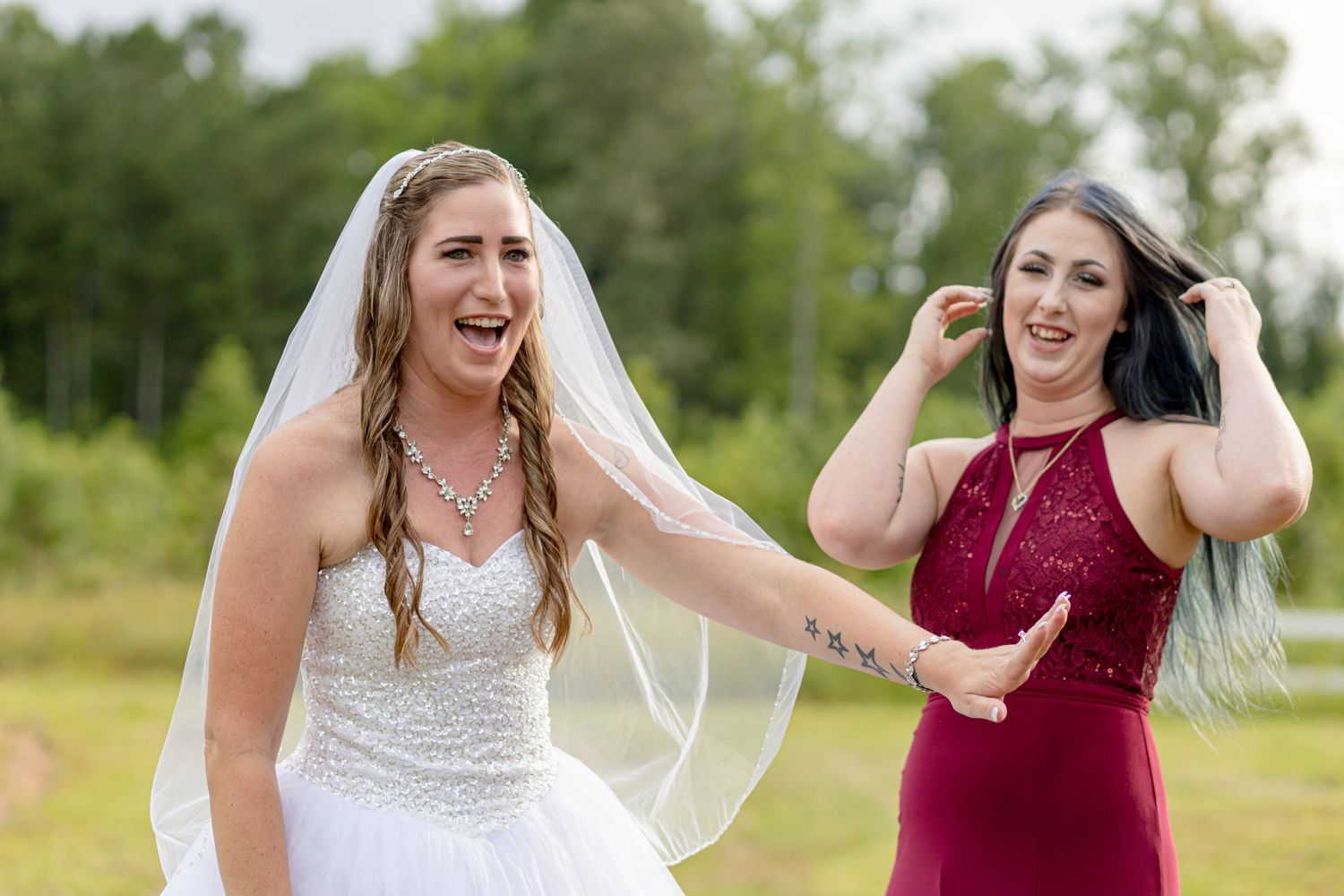 Laughing bride in white gown and veil with bridesmaid in red dress at outdoor wedding.