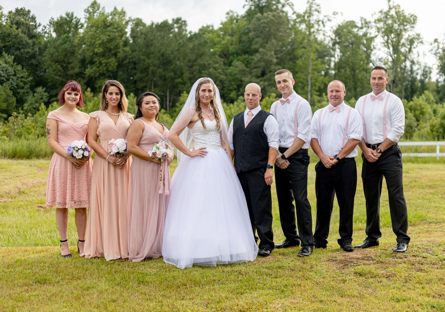 Wedding party posing outdoors with bride in white gown, bridesmaids in blush pink, and groomsmen.
