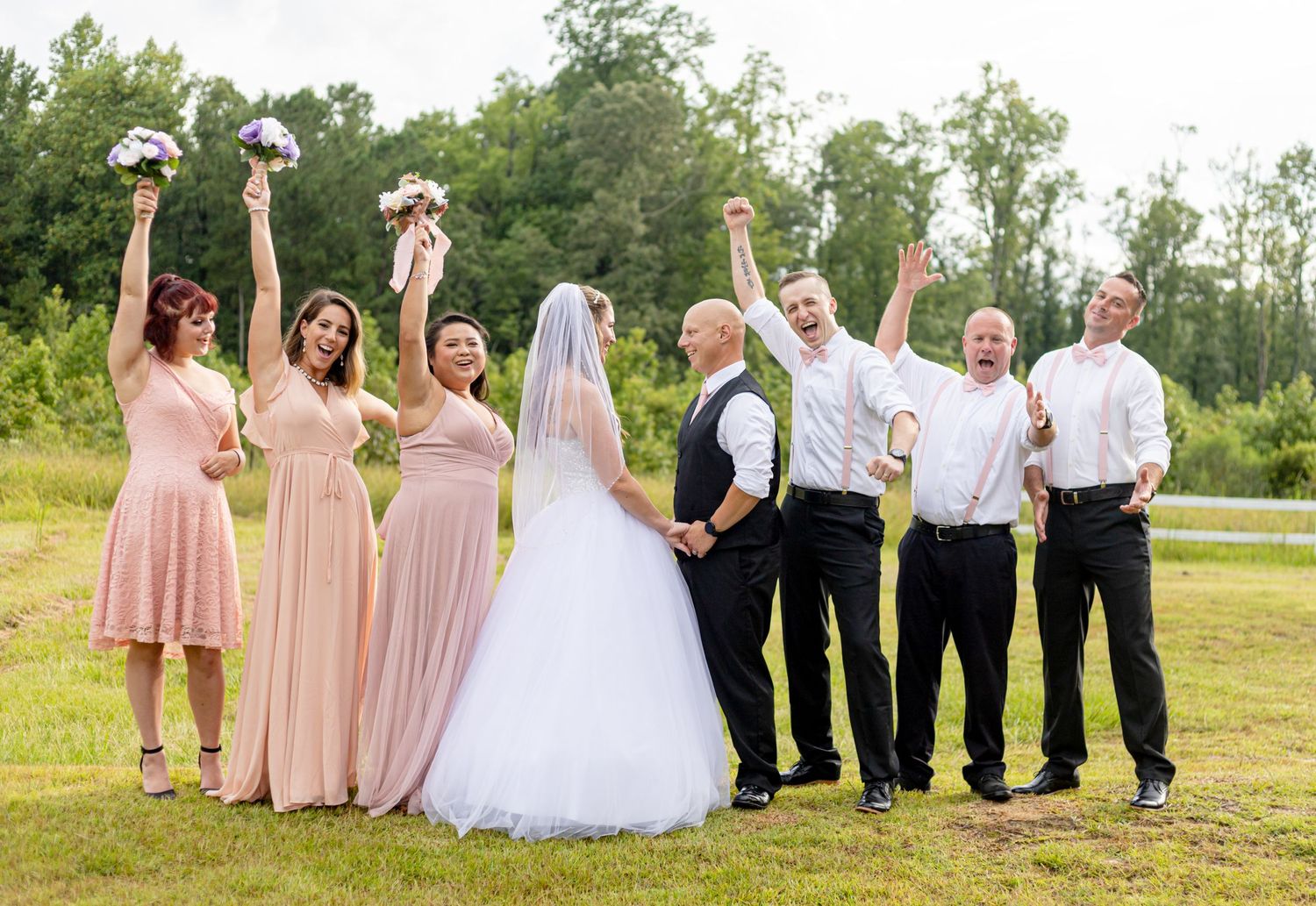 Joyful wedding party celebrating with raised arms in green outdoor setting.