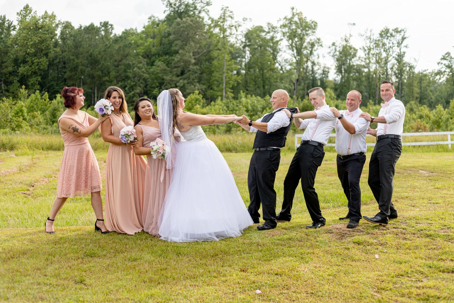 Fun wedding party photo with bridal party forming a chain on green lawn outdoors.