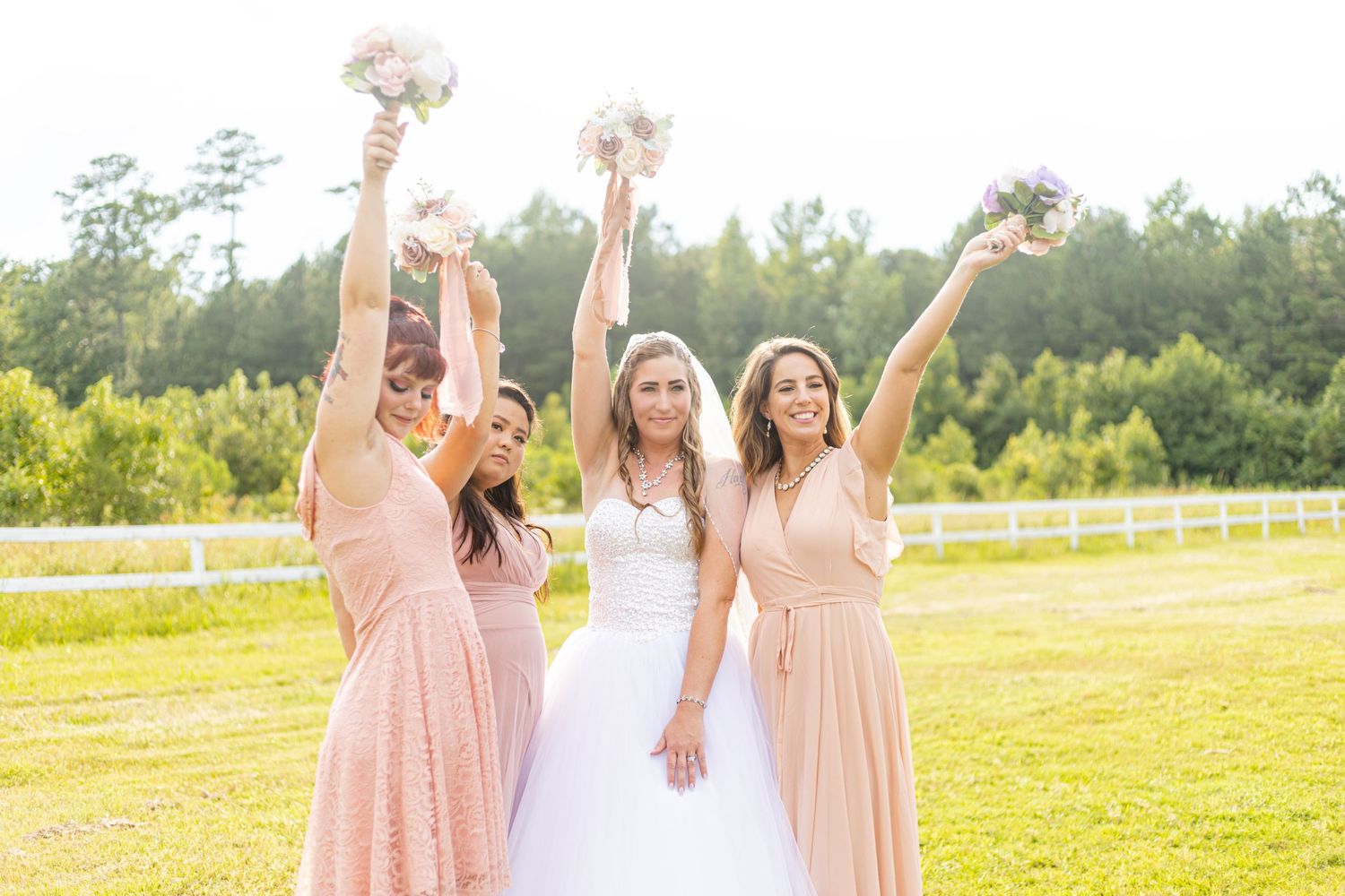 Bride in white gown and three bridesmaids in blush pink raising bouquets joyfully outdoors.
