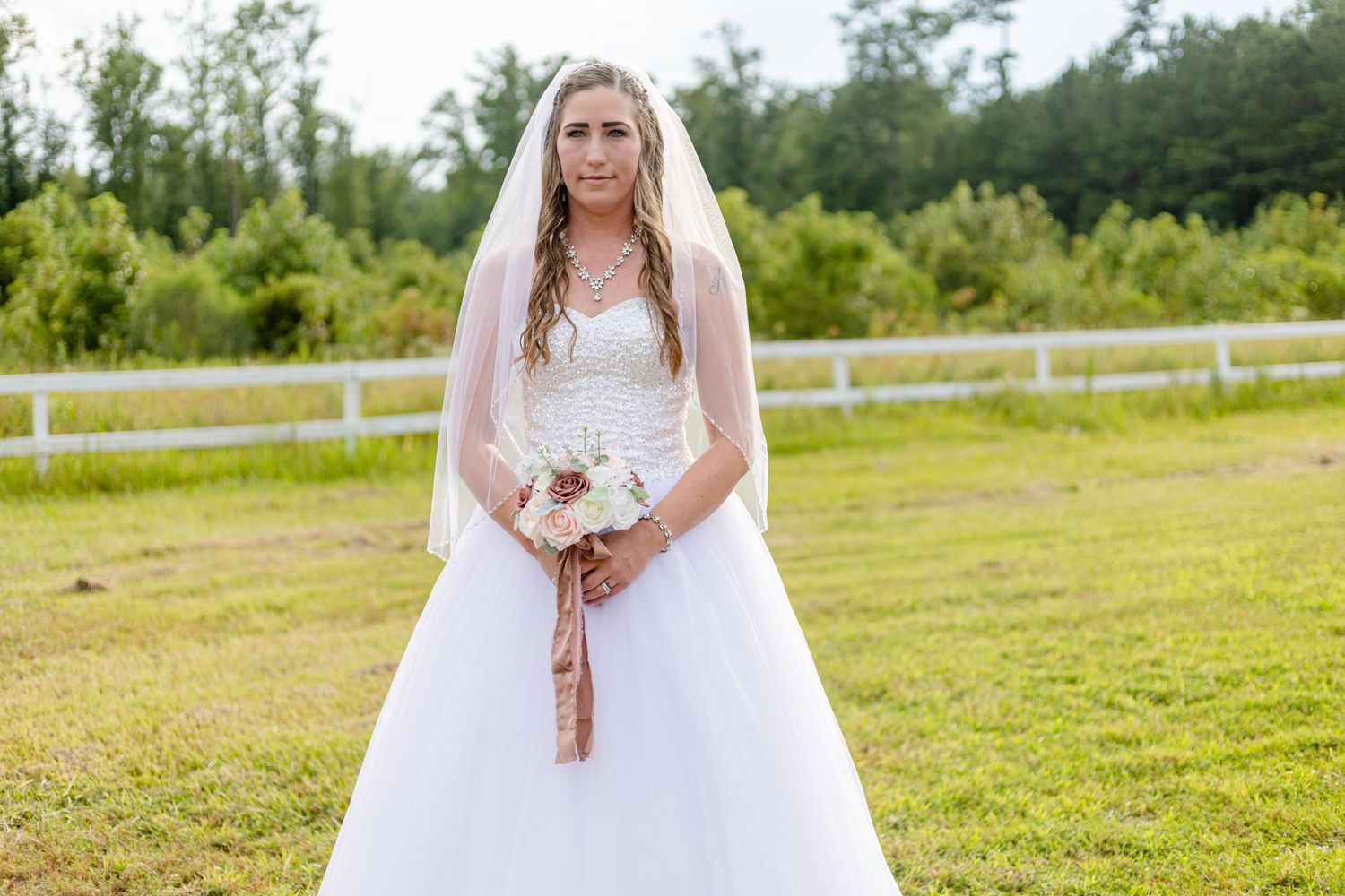 Bride in white ball gown and veil holding bouquet standing in green outdoor setting.