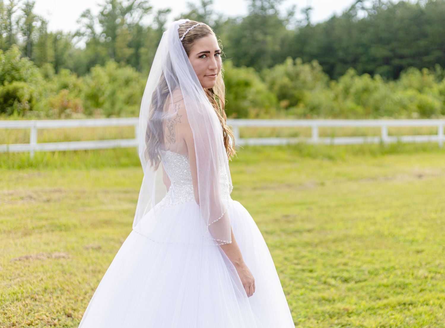Elegant bride in white gown and veil smiling over shoulder in sunny outdoor setting.
