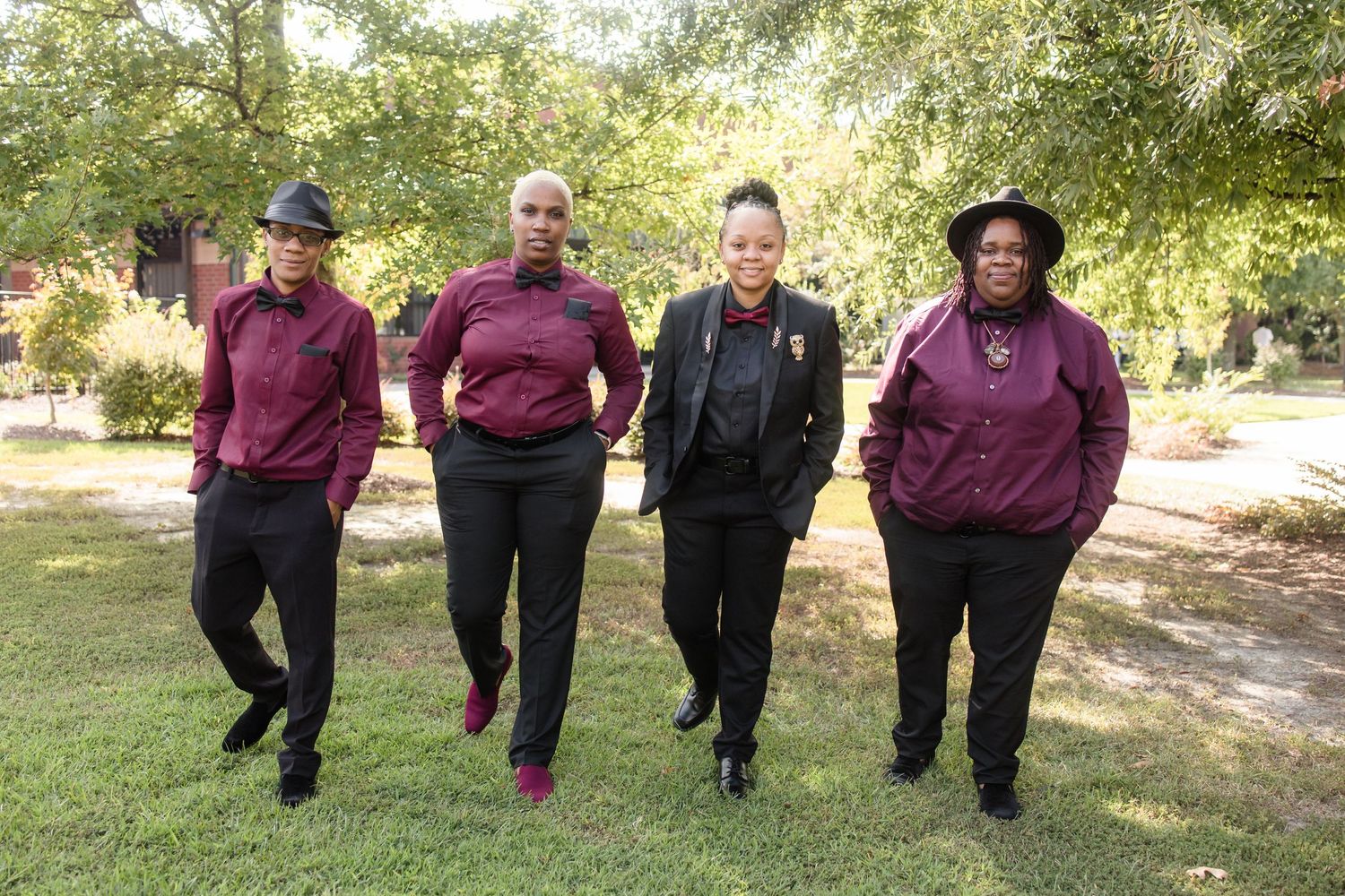 Four women in matching burgundy and black outfits pose together outdoors in a park setting.