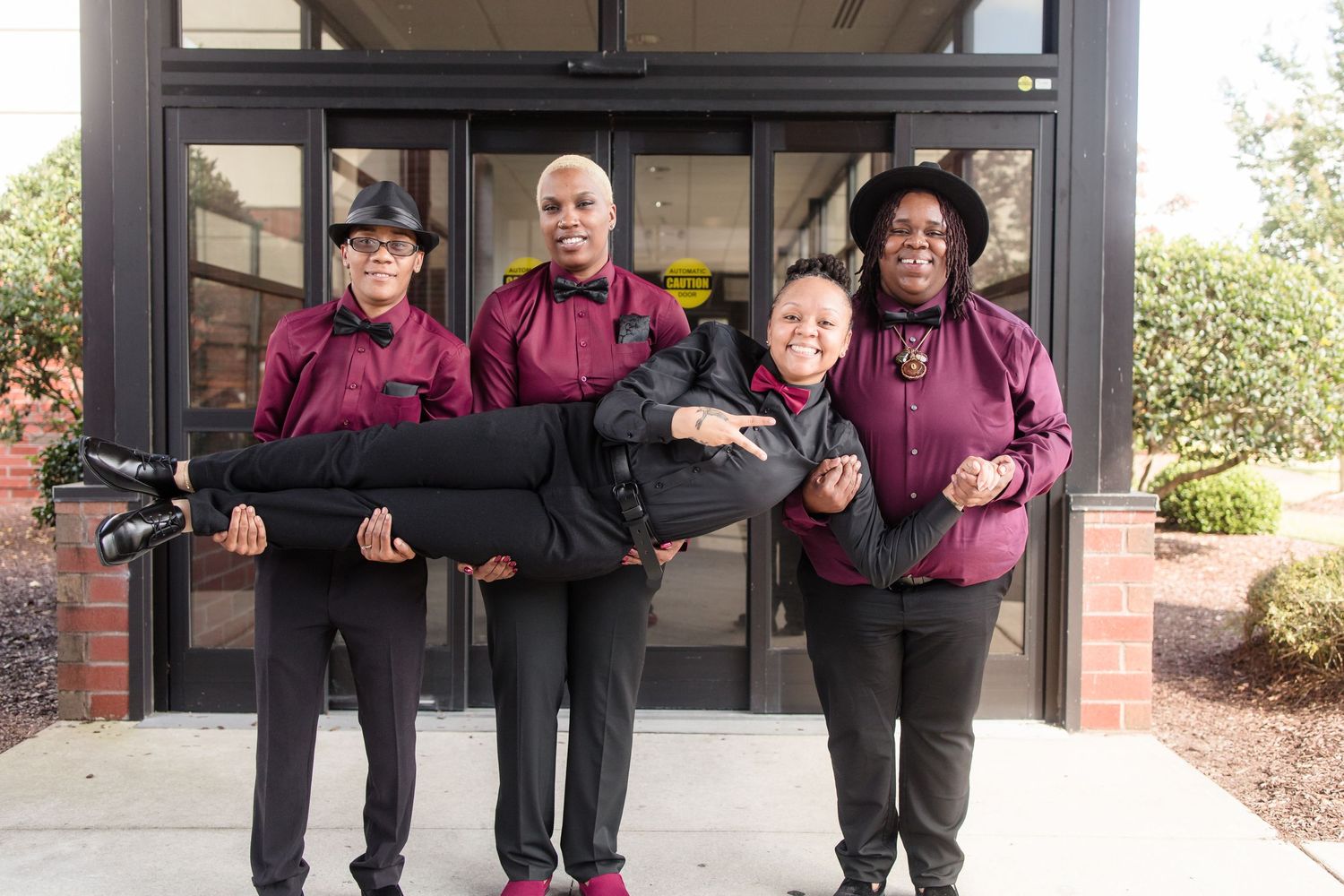 Three women playfully carry a fourth woman in front of a building entrance in burgundy uniforms.
