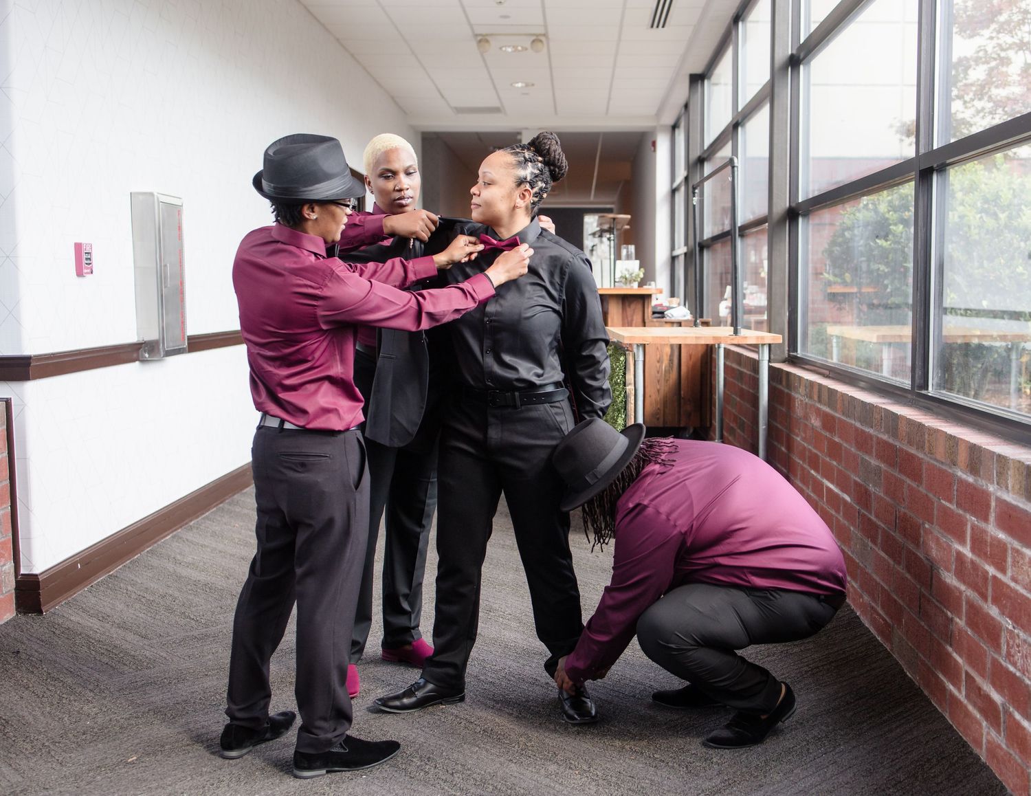 Four women in burgundy and black outfits help each other get dressed in a hallway.