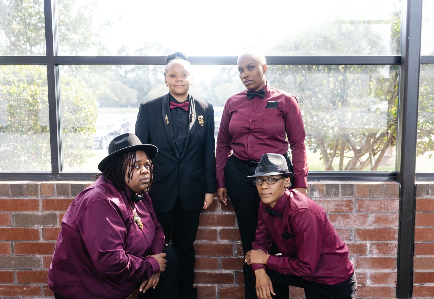 Four women in coordinating burgundy and black wedding attire pose near a brick wall indoors.