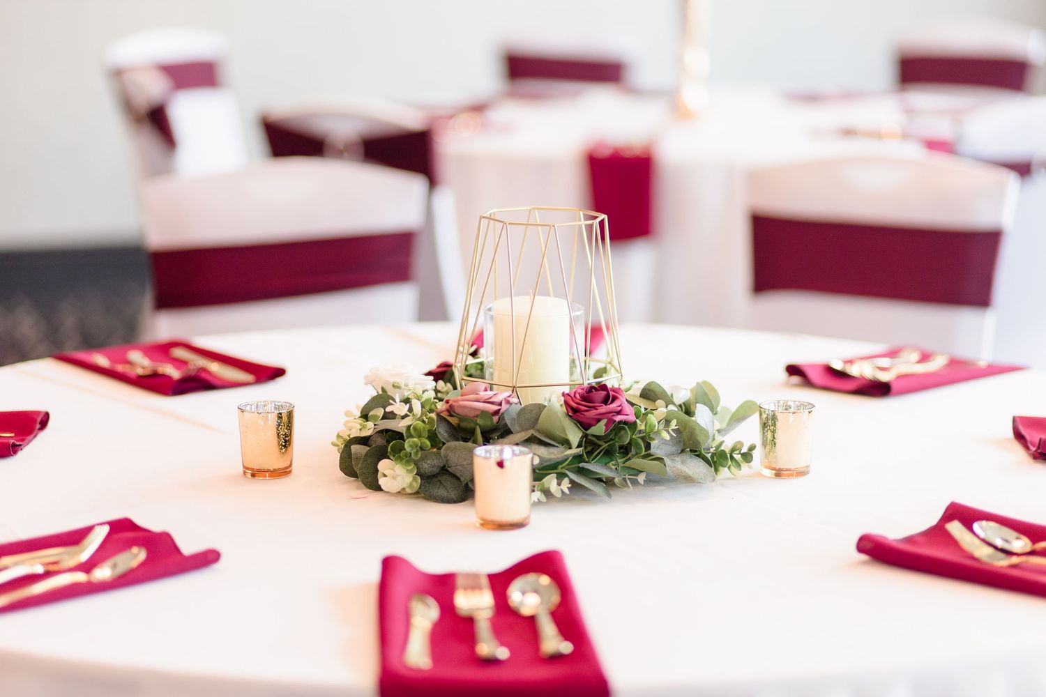 Wedding reception table with geometric candle holder centerpiece, greenery, and burgundy napkins.