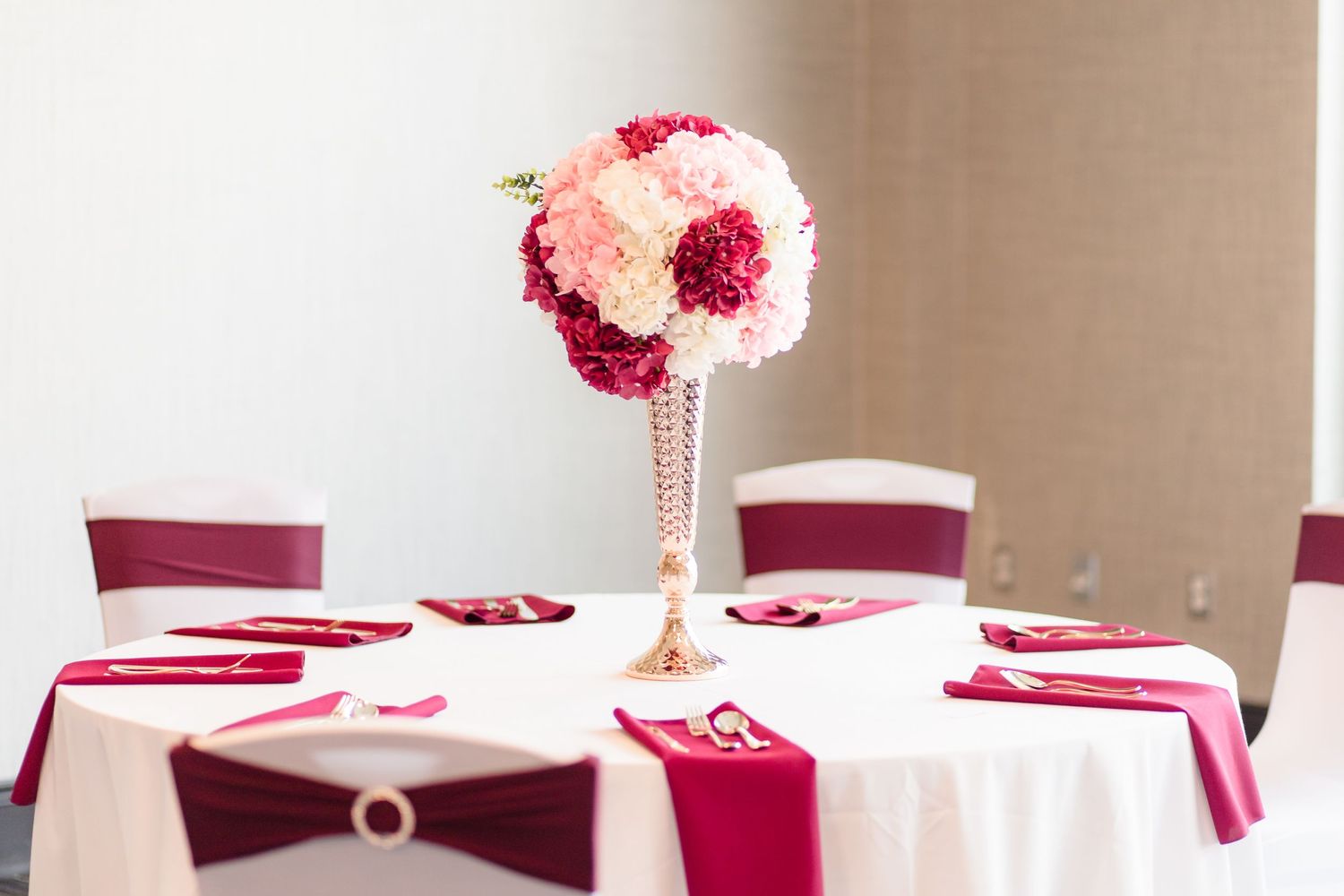 Tall gold vase with pink and burgundy floral arrangement on a white wedding reception table.
