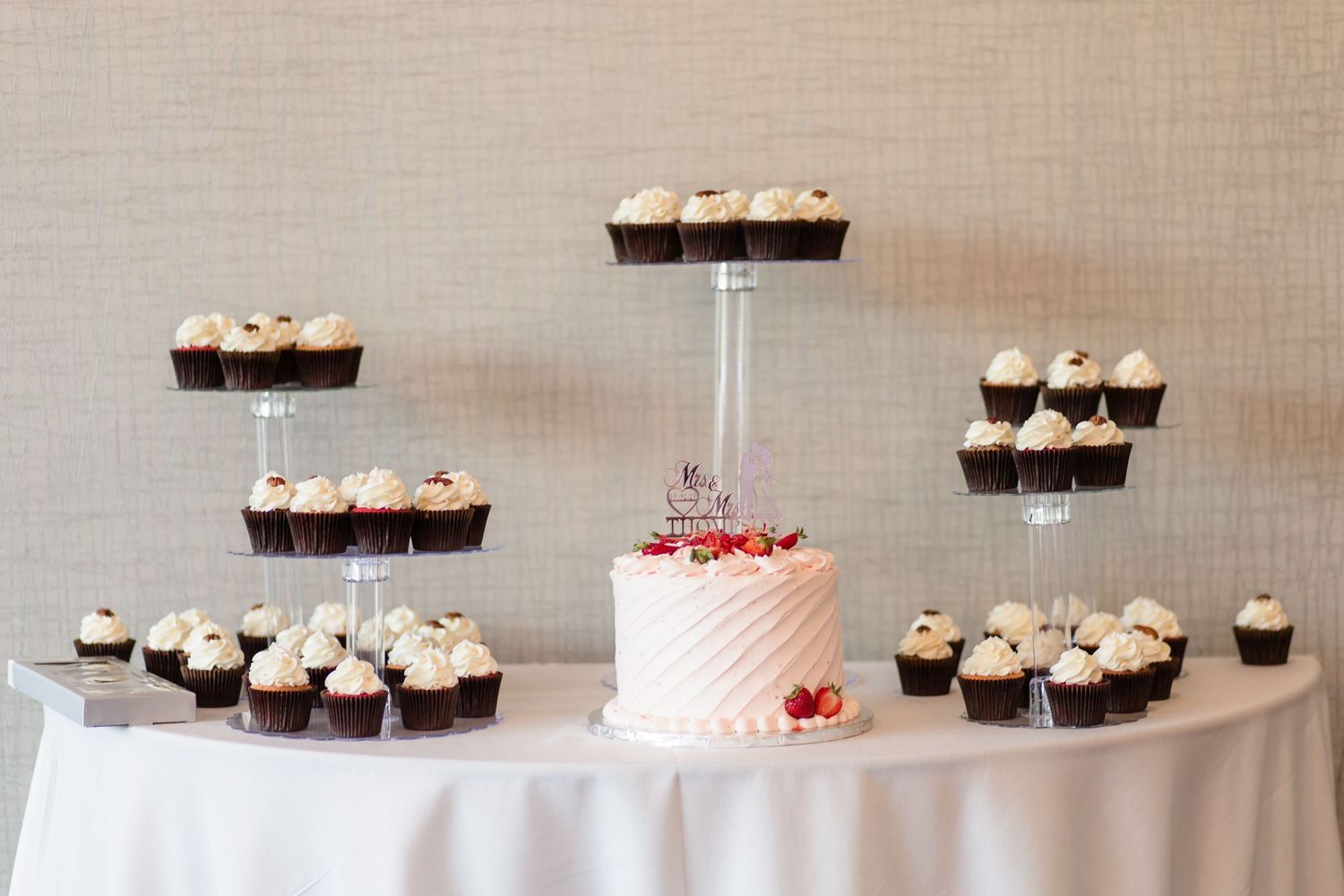 Wedding dessert table featuring a small pink cake surrounded by chocolate cupcakes on tiered stands.