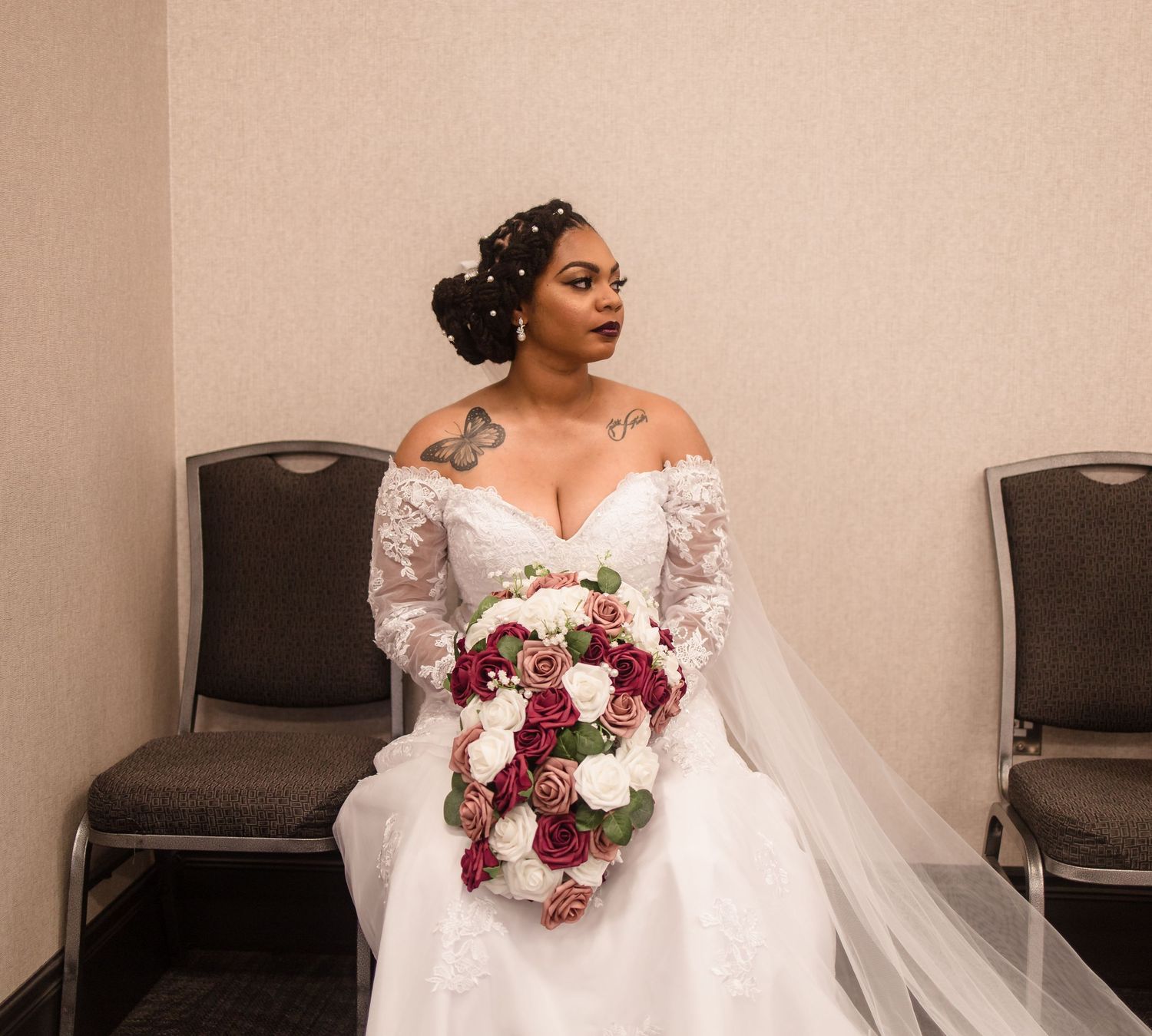 Beautiful bride in off-shoulder lace wedding gown holding burgundy and white rose bouquet while seated.