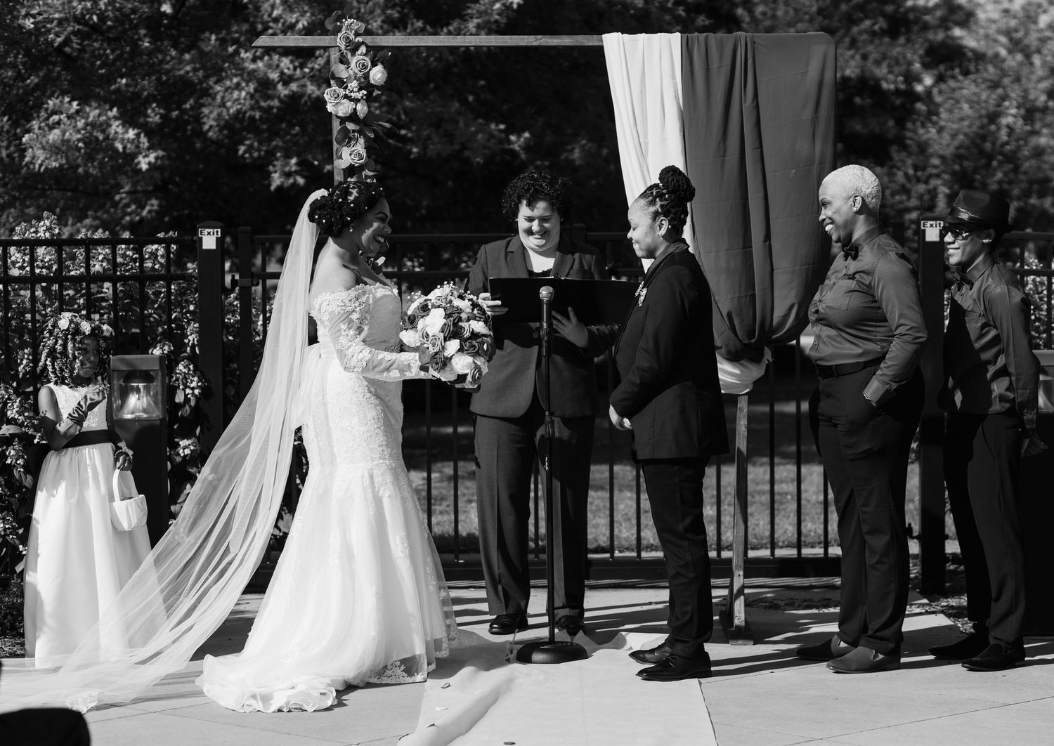 Black and white photo of outdoor wedding ceremony with bride, groom, and officiant at decorated altar.