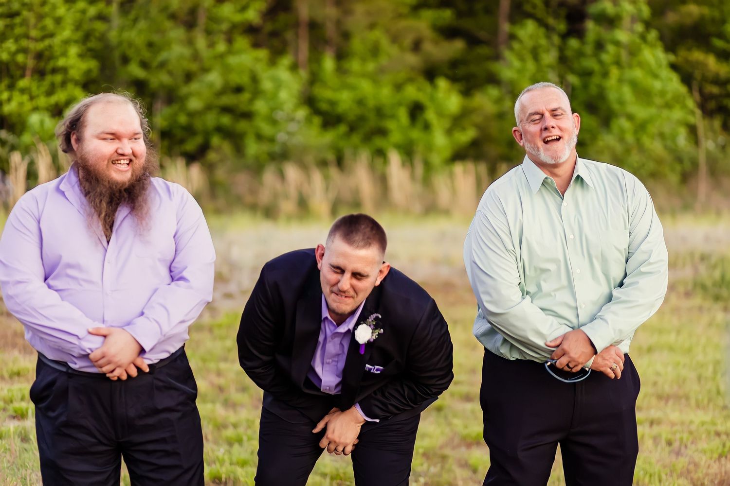 Three men in formal attire laugh together during outdoor wedding celebration in green field.
