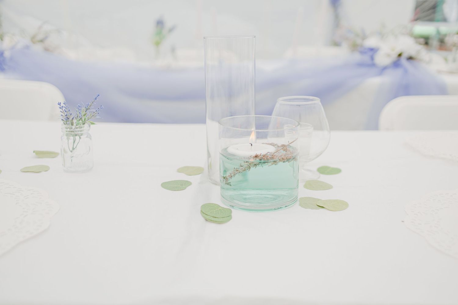 Minimalist wedding table decor with teal candle in glass vase, wine glass, and green leaf confetti on white linen.