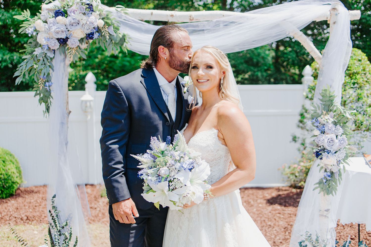 Bride and groom pose under floral arch with blue and white flowers at outdoor wedding.
