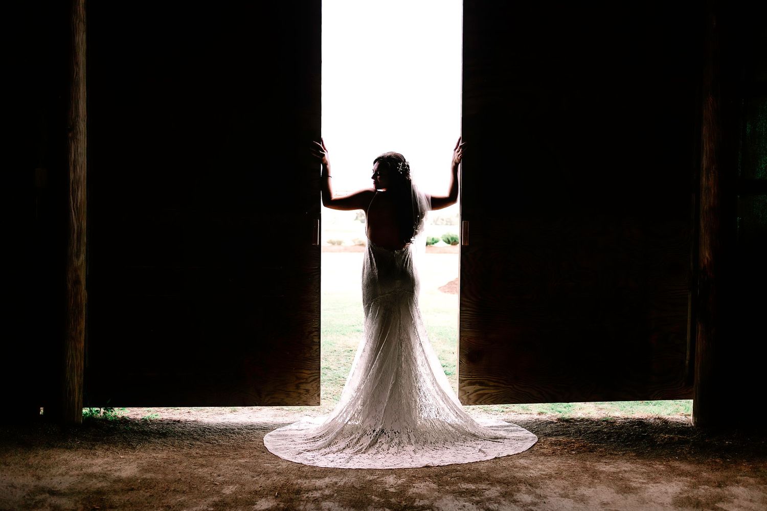 Dramatic silhouette of bride in lace gown with long train standing in open barn doorway.