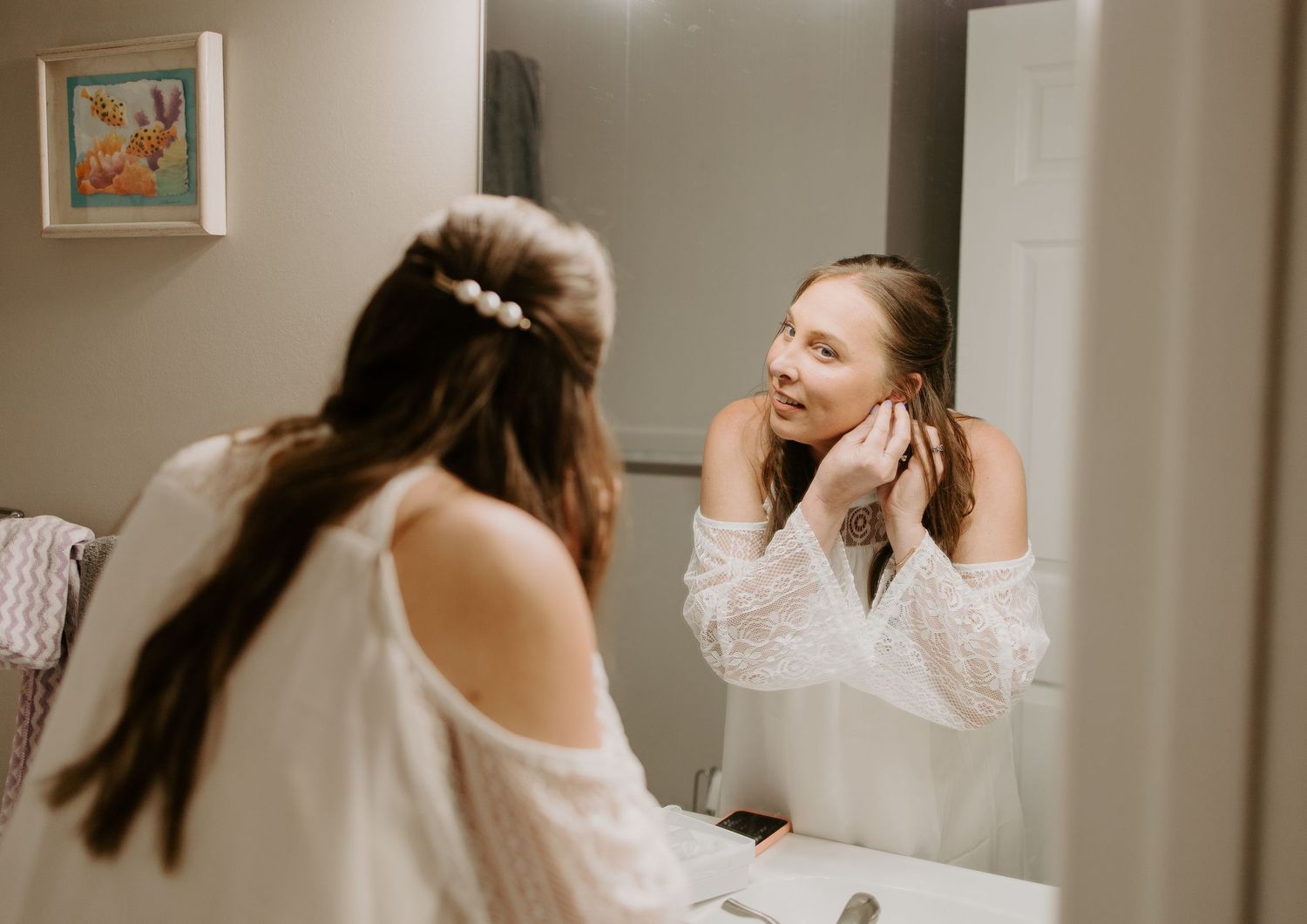 Smiling bride adjusts earrings while getting ready in front of bathroom mirror on wedding day.