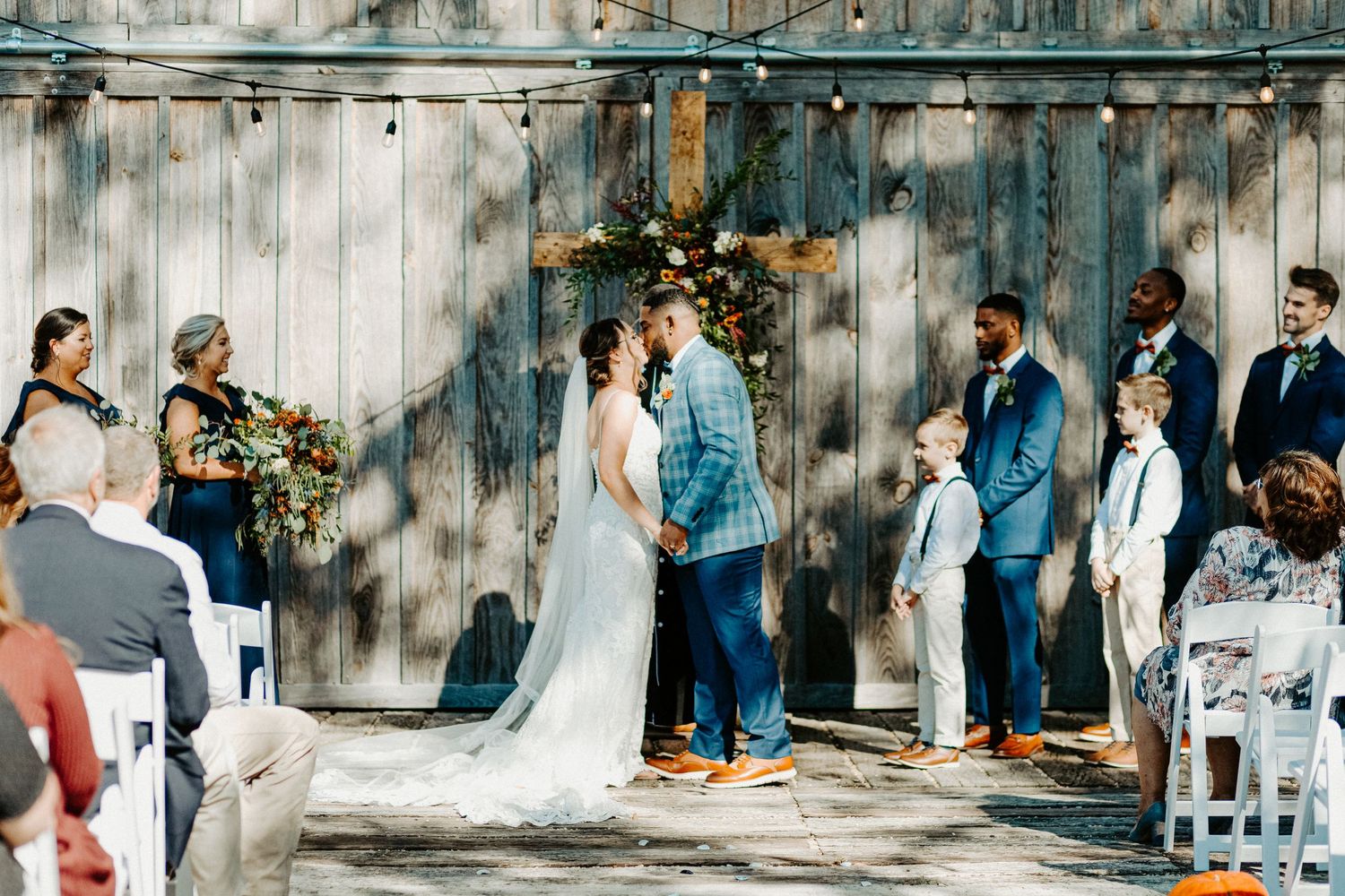 Couple kisses at rustic barn wedding ceremony with wooden backdrop and seated guests.