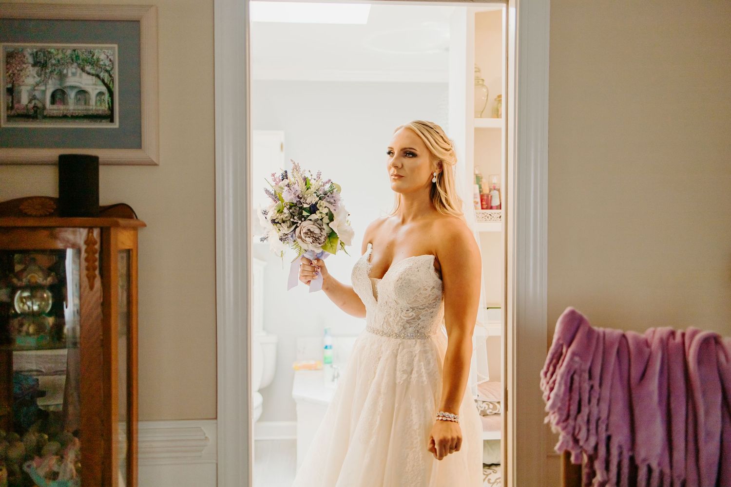 Blonde bride in strapless lace gown holds purple and white bouquet in a doorway.