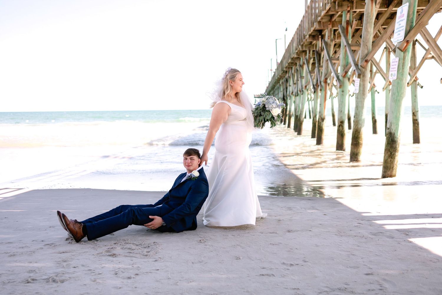 Bride and groom posing on sandy beach near a pier, groom seated casually as bride stands beside him.