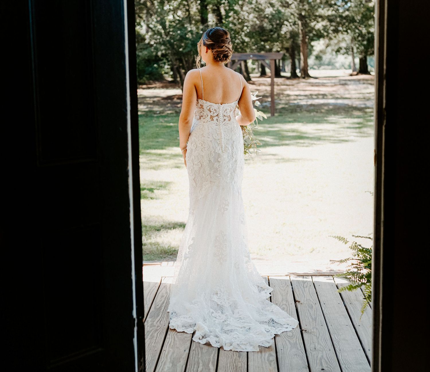 A bride in a fitted lace wedding gown stands on a wooden porch gazing out at a sunlit garden.