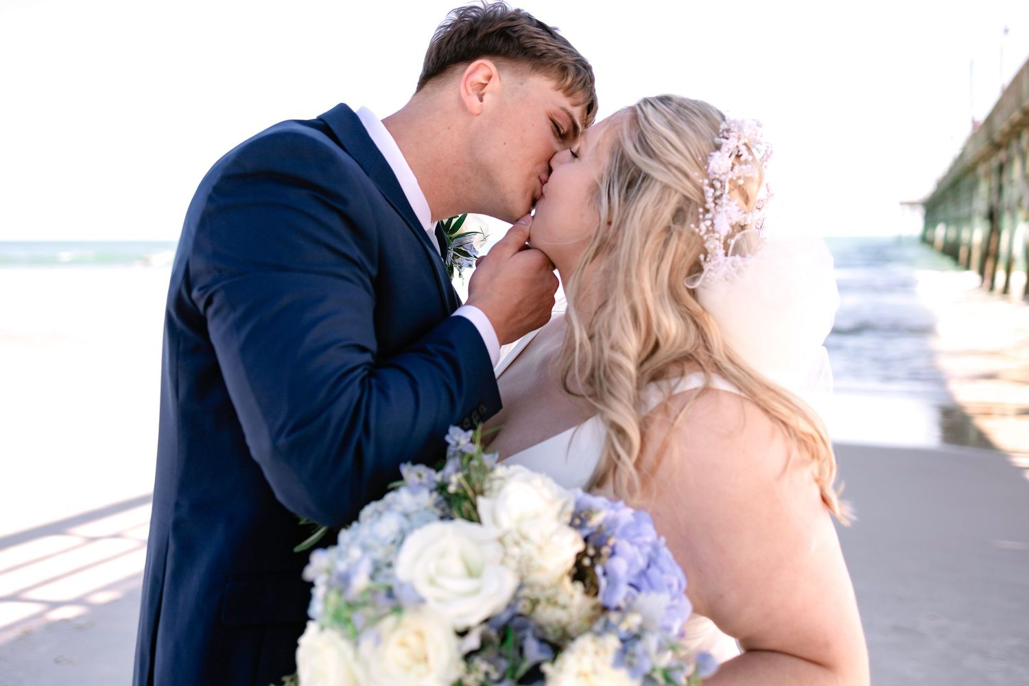 Bride and groom sharing a romantic kiss on the beach near a pier with a white and blue bouquet.