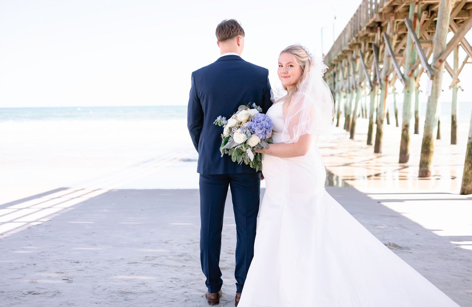 Bride and groom walking hand in hand on the beach near a pier, bride holding a blue and white bouquet.