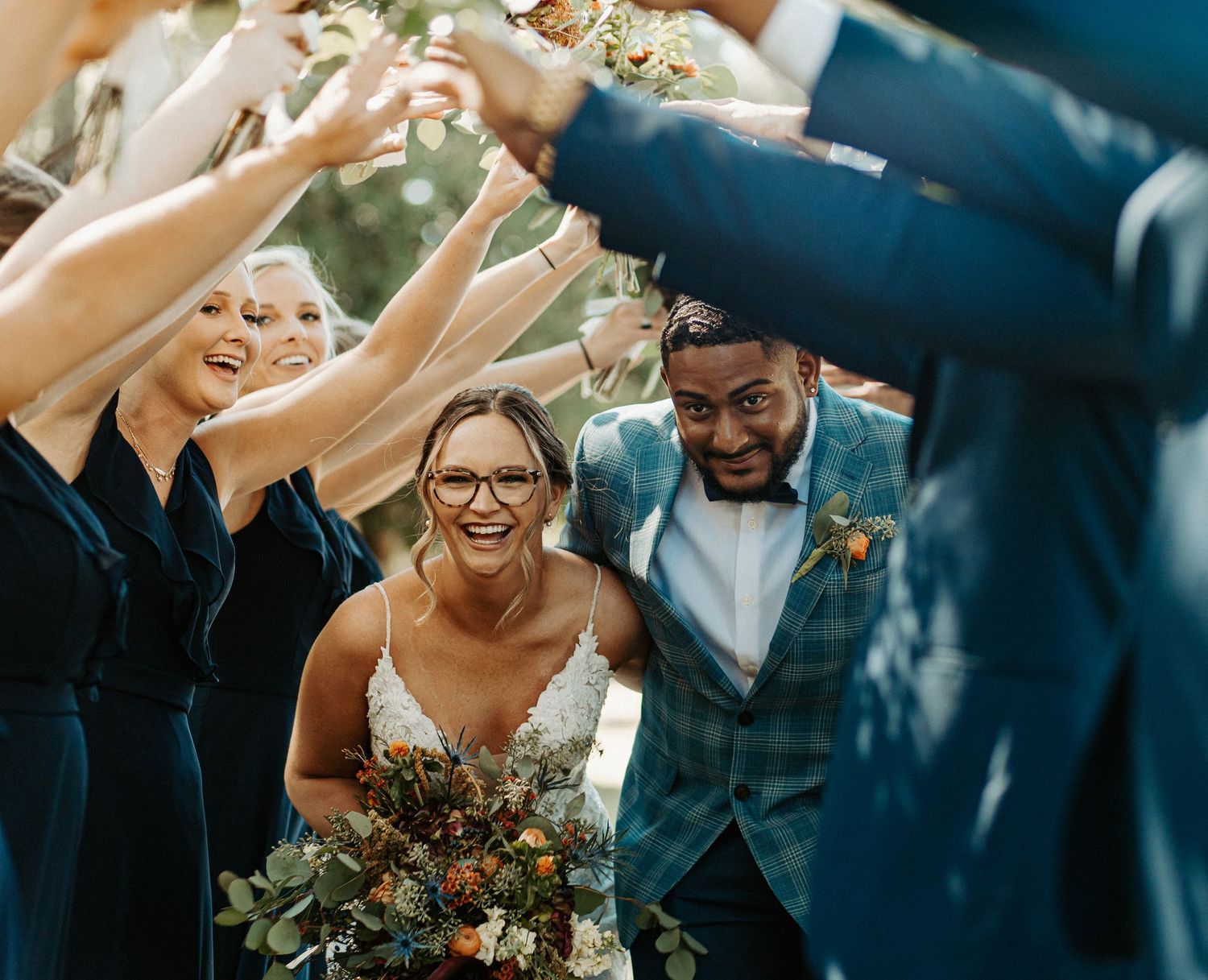Bride and groom walk through tunnel of raised hands formed by smiling wedding party.