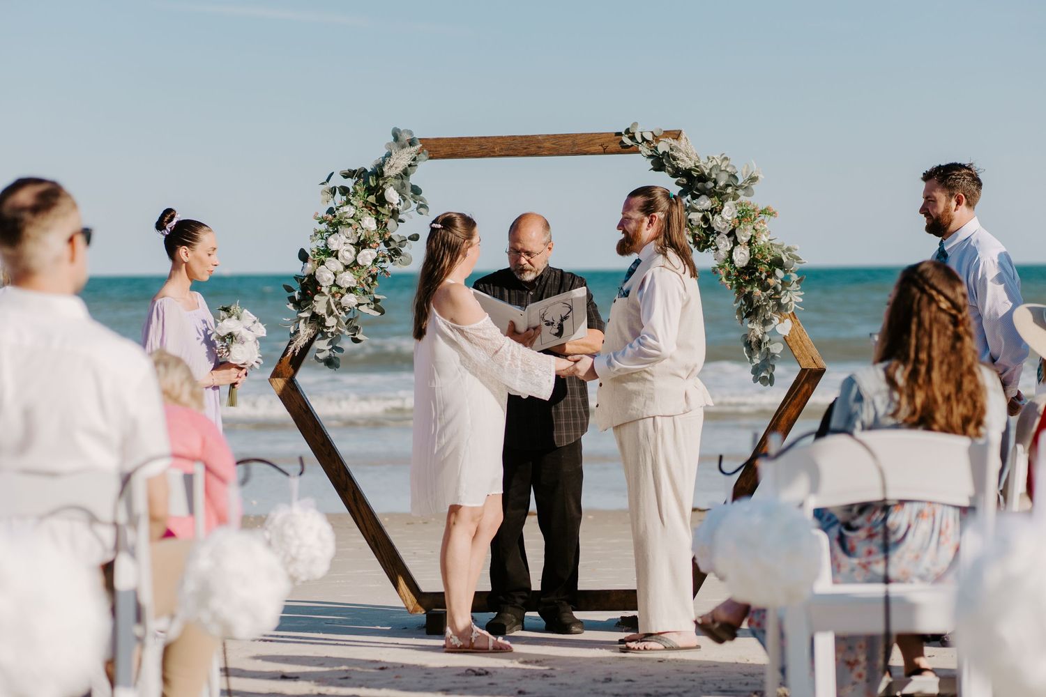 Beach wedding ceremony with couple exchanging vows under a wooden arch adorned with white flowers.