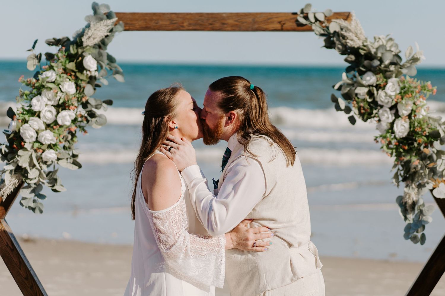 Bride and groom share first kiss under floral hexagonal arch on a sunny beach wedding.
