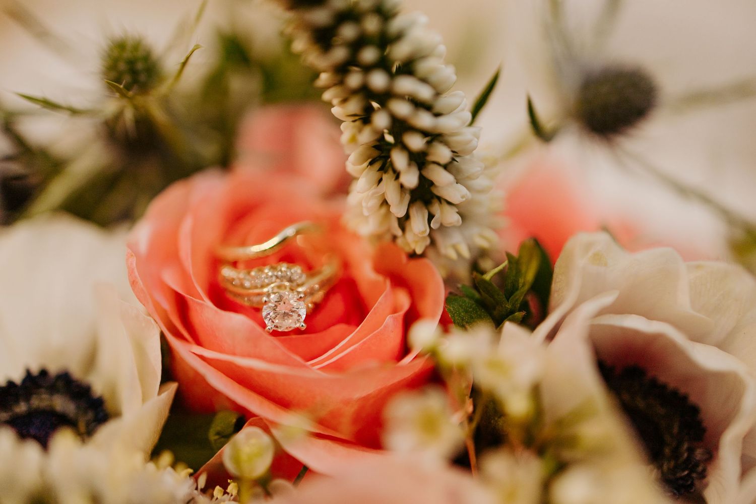 Gold diamond engagement ring resting in a coral pink rose surrounded by a lush wedding bouquet.