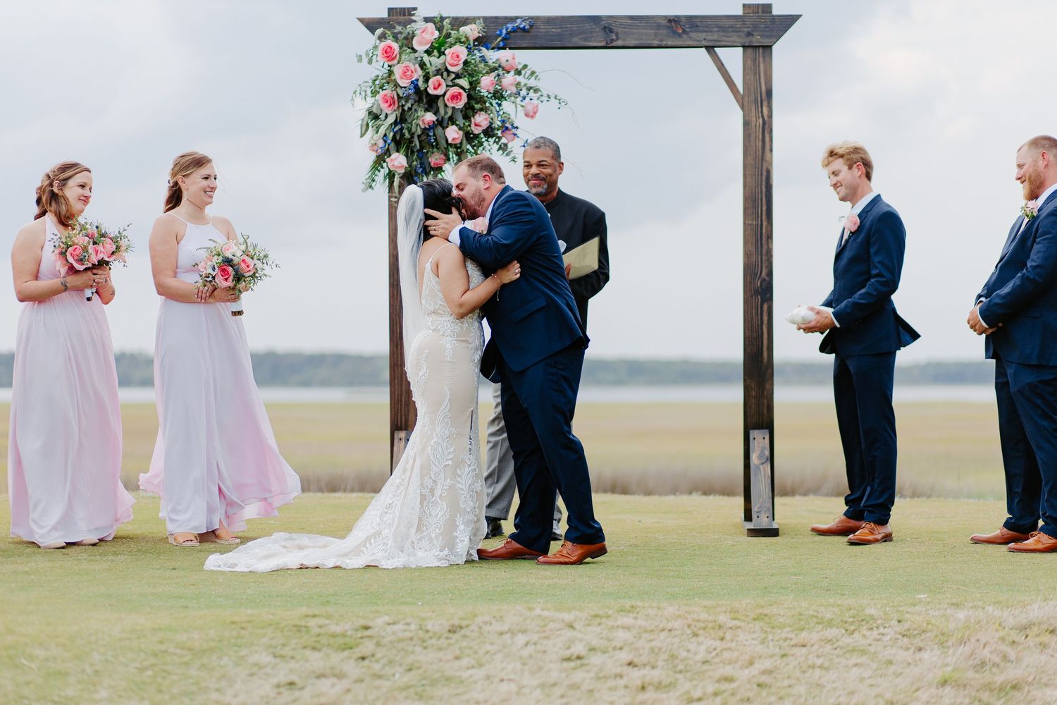 Newlyweds share first kiss at outdoor wooden arch ceremony on open waterfront lawn.