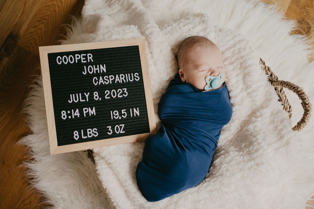 Newborn baby in blue swaddle next to birth announcement board on white rug.