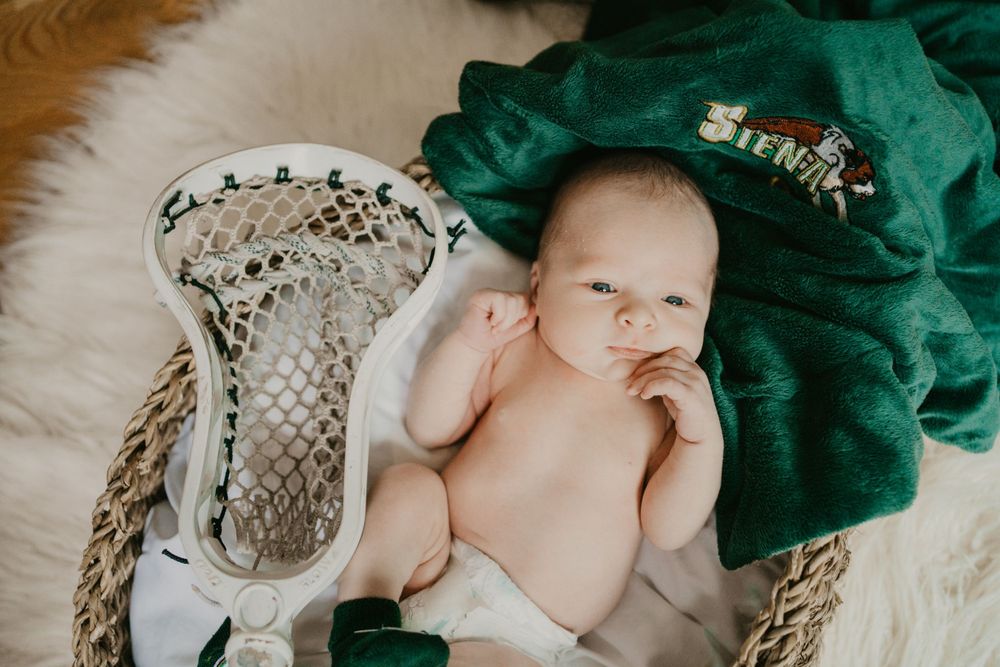 Baby lying next to lacrosse stick and green jersey on white furry backdrop.